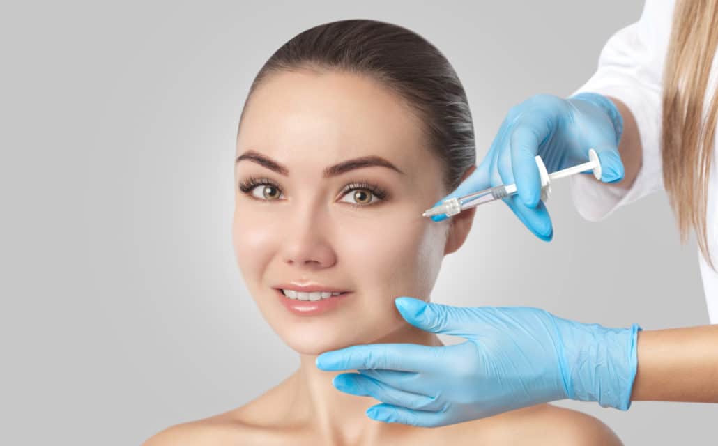 an image of a woman recieving a dermal fillers injection in her cheek. The background is a grey blank canvas, the doctor is wearing blue latex gloves.